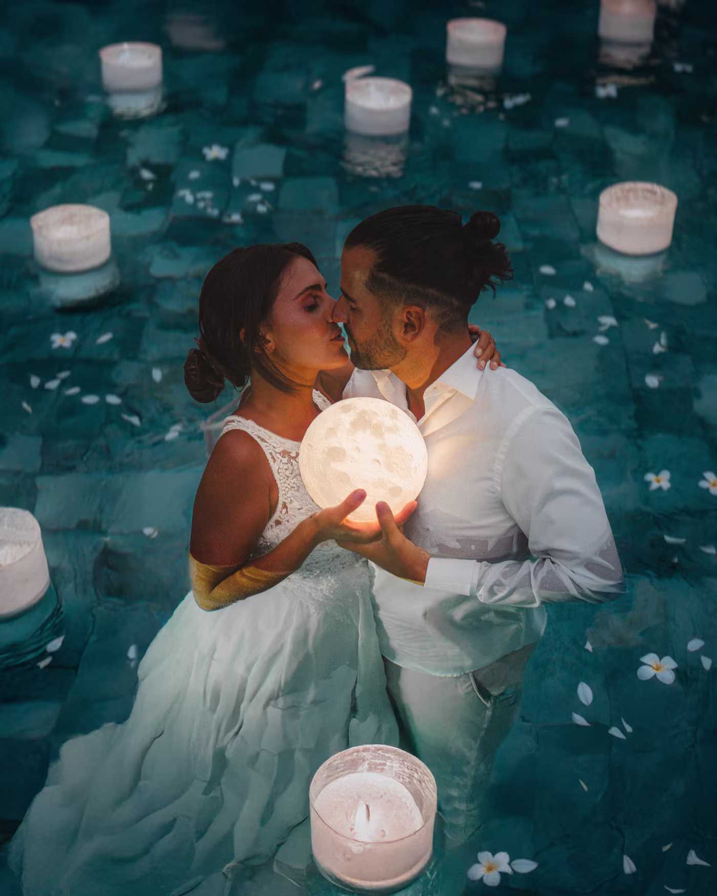 Bride and groom kissing in a pool at night holding a glowing moon lamp among floating candles