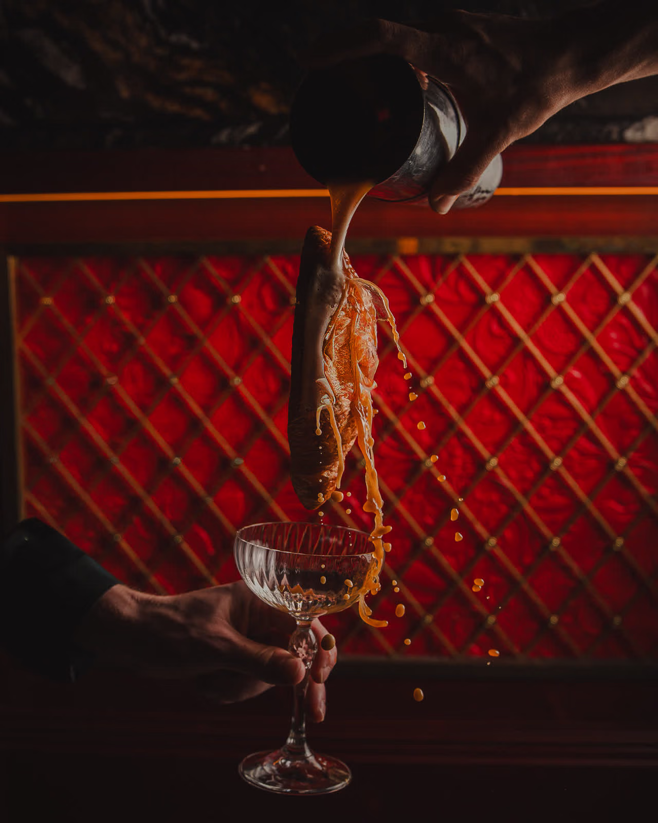 Cocktail poured over a croissant into a coupe glass at a red quilted bar, by Gorka Di Capitan