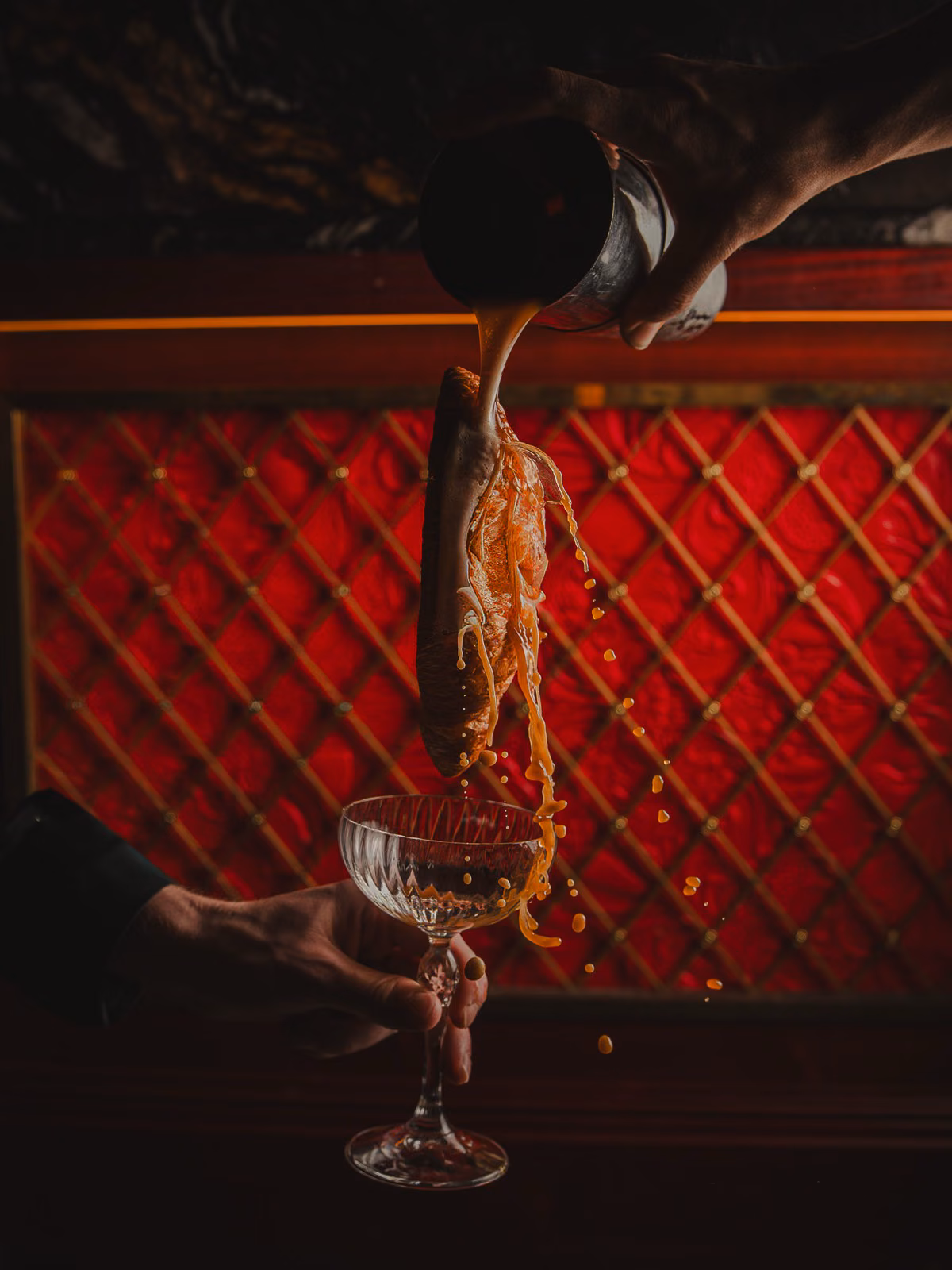 Cocktail poured over a croissant into a coupe glass at a red quilted bar, by Gorka Di Capitan