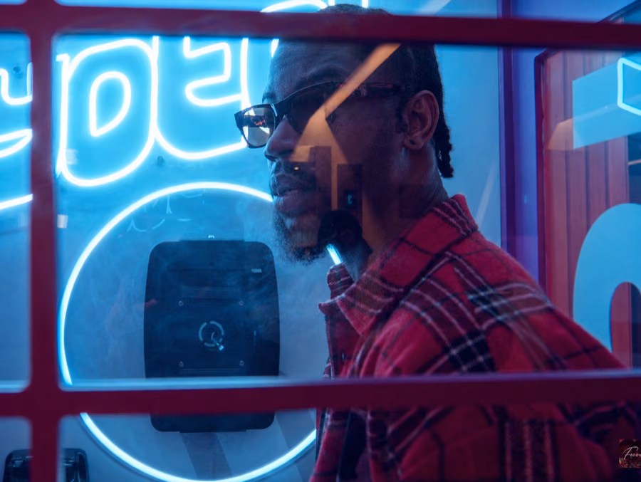Nelson Mvndelv in a red plaid shirt behind neon-lit glass, still from the Morena music video